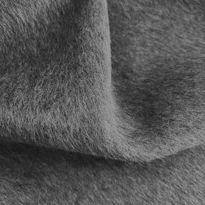 May include: Close-up of a folded section of gray faux fur fabric. The fabric has a soft, fluffy texture with long, fine fibers. The color is a solid, medium gray, with subtle variations in tone due to the light.