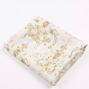 May include: Folded white fabric with a gold floral pattern. The design features ornate, swirling leaves and flowers, creating a classic, elegant look. The fabric is folded neatly, showcasing the detailed pattern.