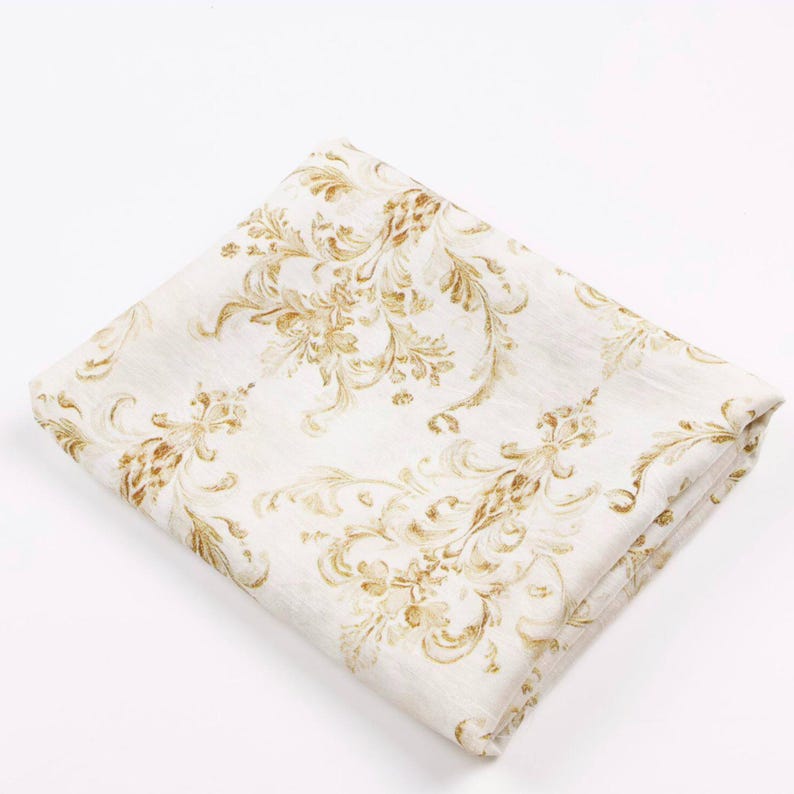 May include: Folded white fabric with a gold floral pattern. The design features ornate, swirling leaves and flowers, creating a classic, elegant look. The fabric is folded neatly, showcasing the detailed pattern.
