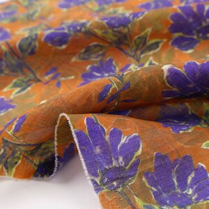 May include: Close-up of a fabric with a floral pattern. The fabric has a burnt orange background with large purple flowers and green leaves. The fabric is folded, showing the texture and design.