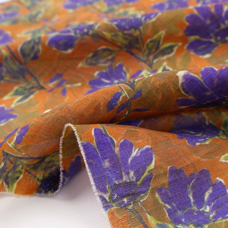 May include: Close-up of a fabric with a floral pattern. The fabric has a burnt orange background with large purple flowers and green leaves. The fabric is folded, showing the texture and design.