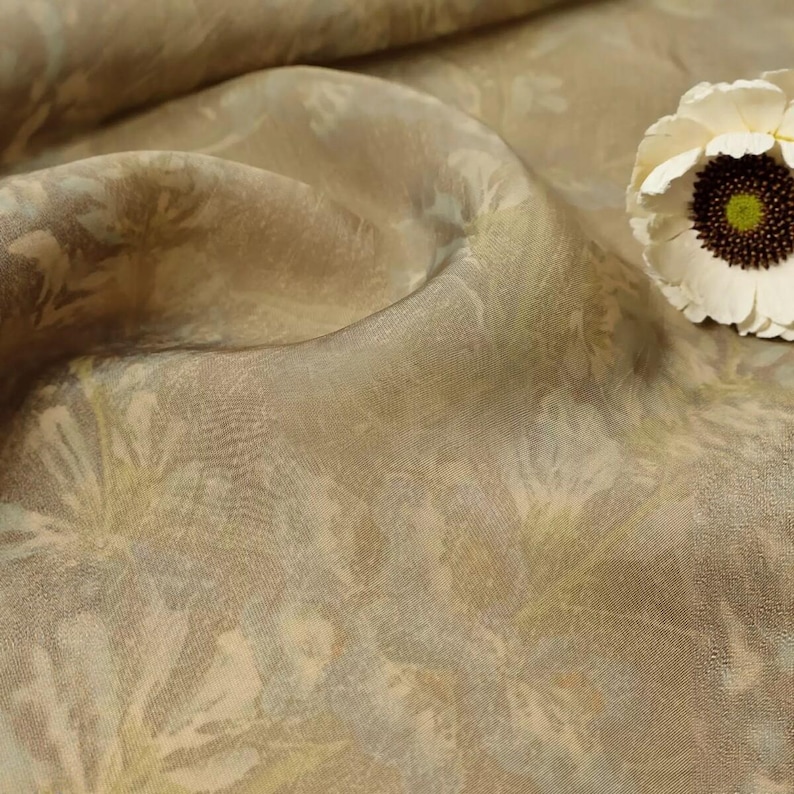 Puede incluir: Una tela transparente de color marr&oacute;n claro con un sutil patr&oacute;n floral tonal, que presenta pliegues y drapeados suaves. Una flor de color crema con un centro marr&oacute;n oscuro y un n&uacute;cleo verde brillante se sit&uacute;a en el lado derecho de la tela.