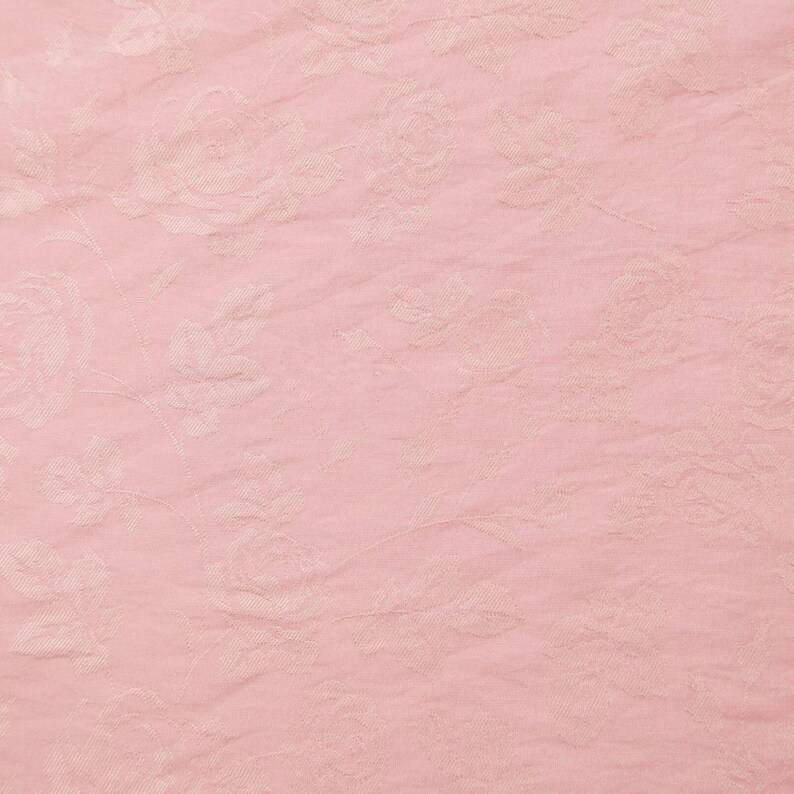 May include: A light pink fabric featuring a subtle, tone-on-tone floral pattern with roses and leaves. The material has a slightly crinkled, textured surface, creating a soft and delicate appearance across the entire visible area of the textile.