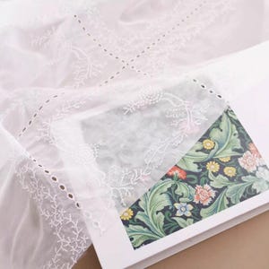 Op de afbeelding: Wit geborduurde stof met bloemen- en bladerpatronen. De stof is gedrapeerd over een boek met een kleurrijk bloemmotief op de kaft. Het borduurwerk heeft delicate ajour.