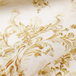 May include: Close-up of a cream-colored fabric with a gold floral damask pattern. The fabric is folded, revealing the texture and detail of the design. The pattern features ornate, swirling leaves and flowers.