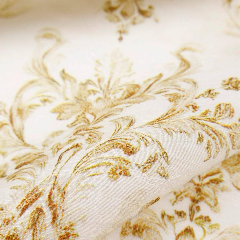 May include: Close-up of a cream-colored fabric with a gold floral damask pattern. The fabric is folded, revealing the texture and detail of the design. The pattern features ornate, swirling leaves and flowers.