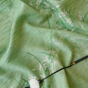 Peut inclure: Un tissu vert clair avec un motif floral de fleurs blanches et de tiges vertes. Le tissu est drap&eacute;, montrant des plis et de la texture. Un cordon noir avec un pendentif blanc est visible.