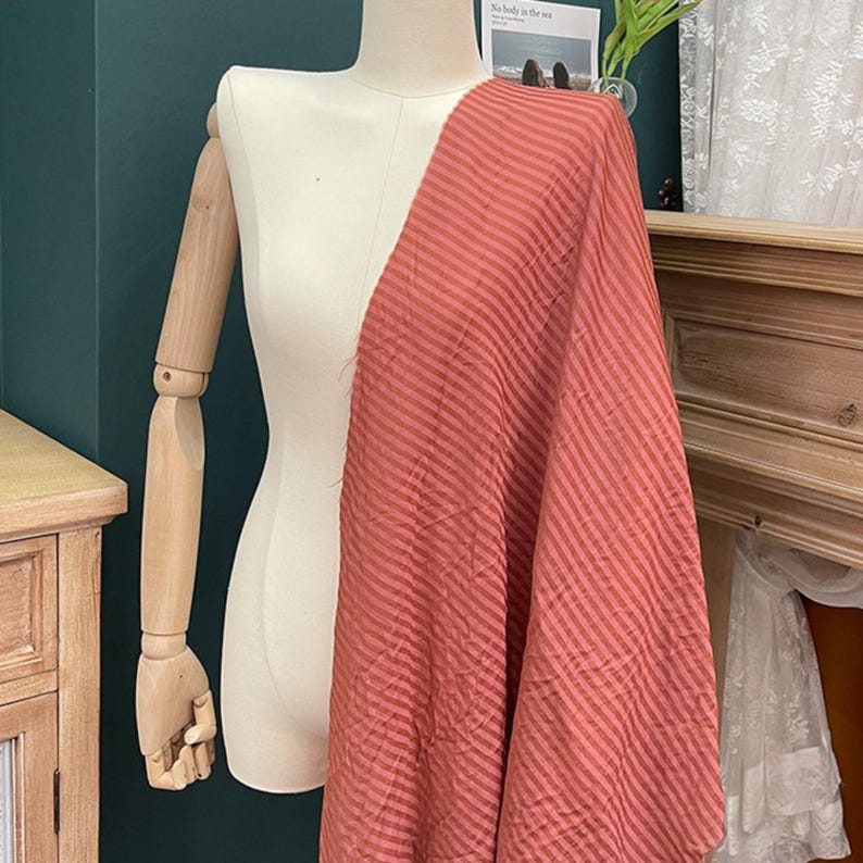 Peut inclure: Une longueur de tissu couleur terre cuite pr&eacute;sentant un motif subtil de rayures diagonales r&eacute;p&eacute;titives. Le textile est drap&eacute; sur un torse de mannequin cr&egrave;me avec des bras en bois articul&eacute;s, mettant en valeur la texture et le tomb&eacute; du mat&eacute;riau.