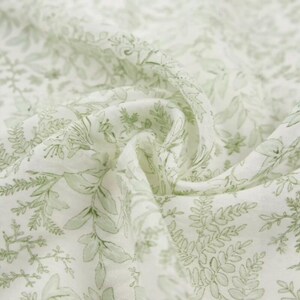 Puede incluir: Primer plano de una tela blanca con un delicado patr&oacute;n de hojas y elementos florales en verde claro. La tela es suave y fluida, con pliegues y arrugas visibles, lo que sugiere un material ligero como el algod&oacute;n o el lino.