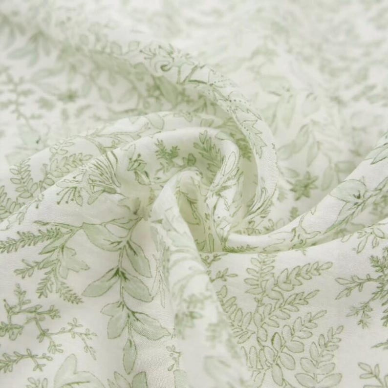Puede incluir: Primer plano de una tela blanca con un delicado patr&oacute;n de hojas y elementos florales en verde claro. La tela es suave y fluida, con pliegues y arrugas visibles, lo que sugiere un material ligero como el algod&oacute;n o el lino.
