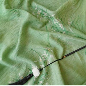 Peut inclure: Un tissu vert clair avec un motif floral de fleurs blanches et de tiges vertes. Une gland noir avec une perle en forme de fleur blanche est attach&eacute;e au tissu.