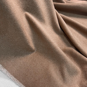 Peut inclure: Gros plan sur un tissu plié de couleur marron chaud et neutre. La matière a un aspect doux et texturé avec des plis et des ombres subtils, suggérant un textile luxueux. Le bord du tissu présente une bordure contrastante de couleur plus claire.
