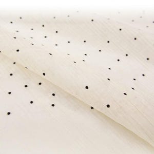 May include: Off-white fabric with a delicate texture and scattered black polka dots. The fabric is folded, creating soft shadows and highlighting the material's drape. This textile is suitable for various sewing and crafting projects.