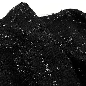 May include: A black fabric with a textured, woven tweed-like surface featuring scattered, reflective sequins throughout. The material has a dense, irregular weave pattern with small, shiny circular embellishments that catch the light across the dark surface.