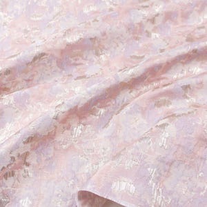 May include: A light pink and lavender fabric featuring a woven floral jacquard pattern with a subtle metallic sheen. The material is draped in soft folds, highlighting the intricate texture and the interplay of the pastel colors across the surface.