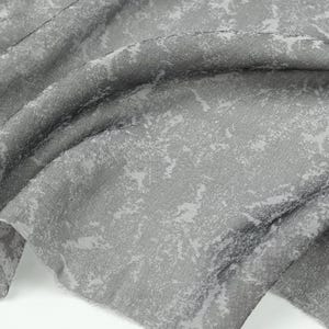 May include: Close-up of a gray fabric with a textured surface and a mottled pattern. The fabric has a soft, flowing drape, with light gray and white accents. The material appears to be a lightweight textile, suitable for apparel or home decor.