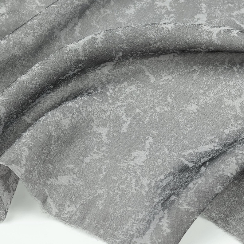 May include: Close-up of a gray fabric with a textured surface and a mottled pattern. The fabric has a soft, flowing drape, with light gray and white accents. The material appears to be a lightweight textile, suitable for apparel or home decor.