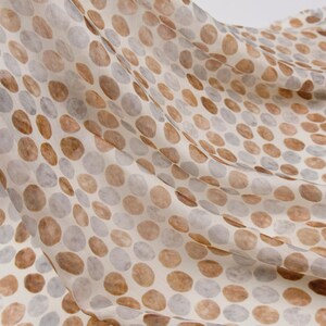 May include: A flowing fabric with a pattern of large, irregular circles in shades of brown and gray against a cream background. The fabric appears soft and lightweight, with visible folds and movement, suggesting it is draped.