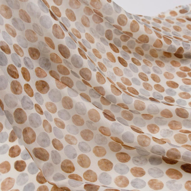 May include: A flowing fabric with a pattern of large, irregular circles in shades of brown and gray against a cream background. The fabric appears soft and lightweight, with visible folds and movement, suggesting it is draped.