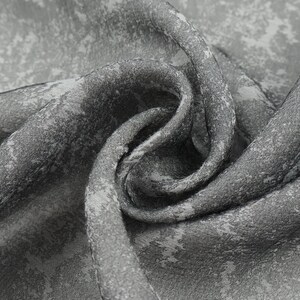 May include: Close-up of a gray fabric with a mottled pattern. The fabric is folded and draped, revealing a textured surface with variations in tone. The design appears to be a mix of light and dark gray hues.