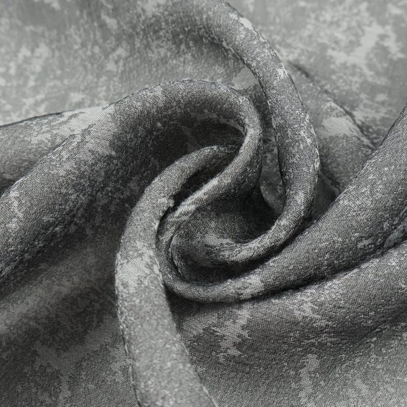 May include: Close-up of a gray fabric with a mottled pattern. The fabric is folded and draped, revealing a textured surface with variations in tone. The design appears to be a mix of light and dark gray hues.