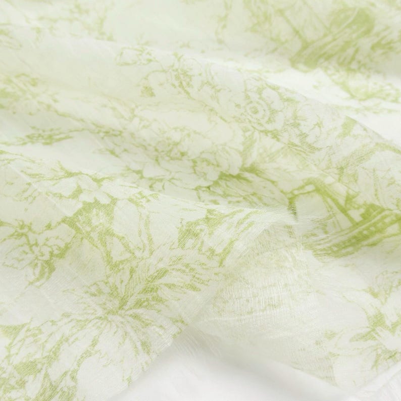 May include: Close-up of a white fabric with a delicate, repeating pattern in shades of green. The design features floral and architectural elements, creating a soft, textured appearance. The fabric is slightly wrinkled, suggesting a lightweight material.