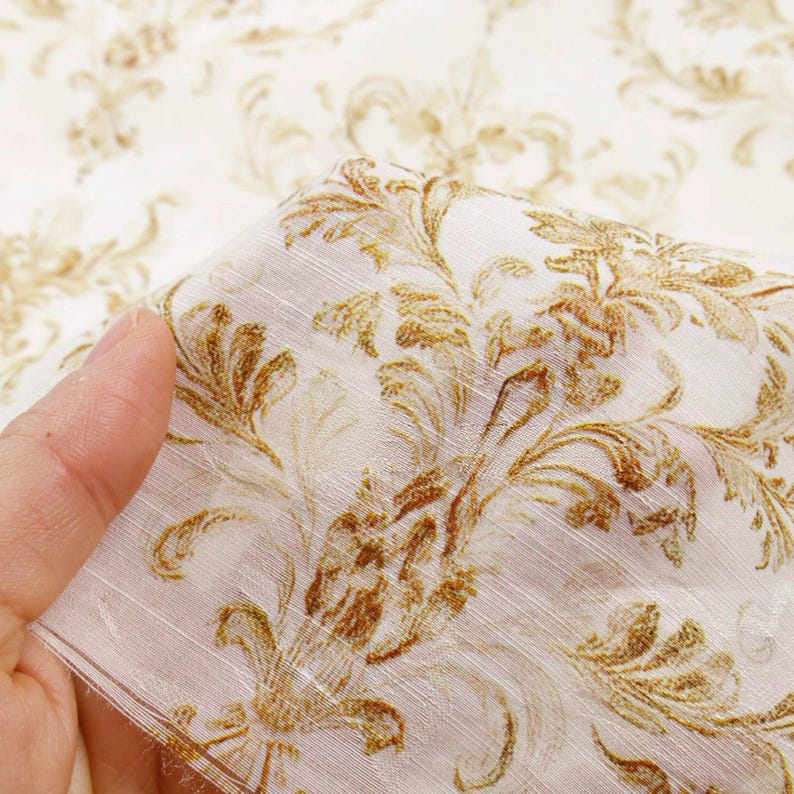 May include: Close-up of a fabric with a cream-colored background and a repeating pattern of gold-colored floral designs. The fabric is folded, revealing its texture and the intricate details of the pattern.