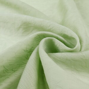 May include: Close-up of a light green fabric with a subtle floral pattern. The fabric is draped, showing folds and textures. The color is a soft, muted green, and the material appears to be lightweight.
