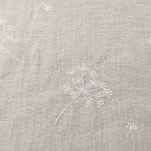 May include: Close-up of a light gray fabric with white embroidered floral patterns. The design features delicate, stylized flowers with multiple small, beaded details. The fabric has a subtle, textured appearance.