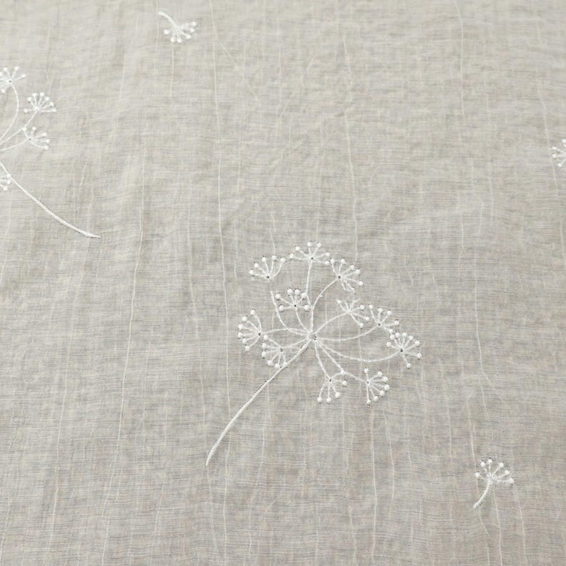 May include: Close-up of a light gray fabric with white embroidered floral patterns. The design features delicate, stylized flowers with multiple small, beaded details. The fabric has a subtle, textured appearance.