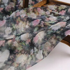 Pu&ograve; includere: Un tessuto trasparente con motivo floreale. Il tessuto presenta uno sfondo scuro con un mix di fiori rosa, bianchi e verdi. Il tessuto &egrave; drappeggiato su una struttura in legno, mostrando la sua texture fluida.