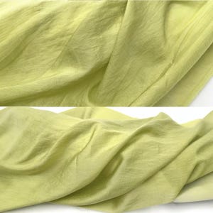 May include: A light lime green fabric with a fine, semi-sheer texture and a subtle horizontal weave pattern. The material is draped in soft, flowing folds, showcasing its lightweight quality and consistent, vibrant yellow-green color throughout.