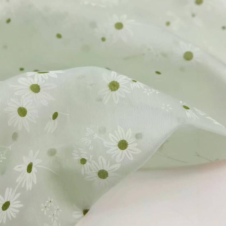 May include: A sheer, light sage green fabric featuring a repeating pattern of white daisy flowers with olive green centers. The material has a delicate, semi-transparent quality with soft folds that create depth and texture across the surface.