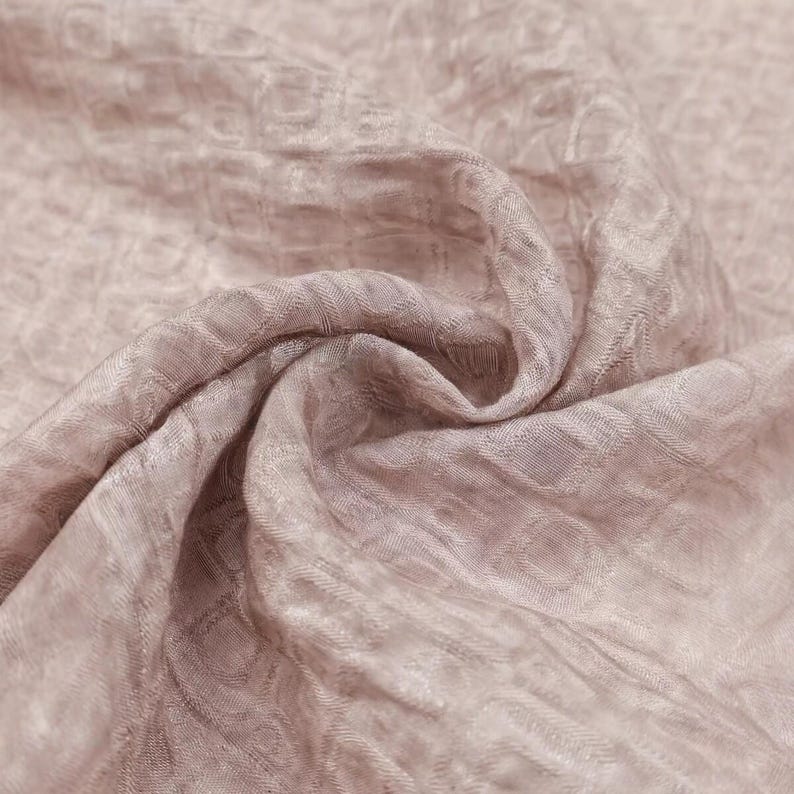 May include: Close-up of a soft, flowing fabric in a muted mauve color. The textile has a subtle, repeating circular pattern and a slightly textured surface, creating a delicate and elegant appearance. The fabric is draped, showing folds and shadows.