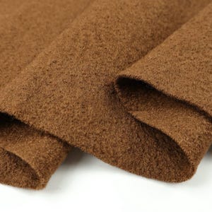 May include: A piece of brown fabric with a dense, fuzzy, and textured surface. The material is folded over itself, creating soft curves and shadows that highlight the consistent, deep brown color and the fibrous, matte finish of the textile.