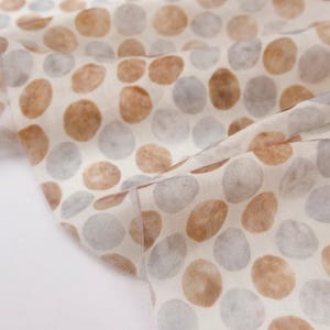 May include: Close-up of a fabric with a repeating pattern of large, irregular circles. The circles are in shades of brown and gray, scattered across a cream-colored background. The fabric is folded, showing the texture and drape of the material.