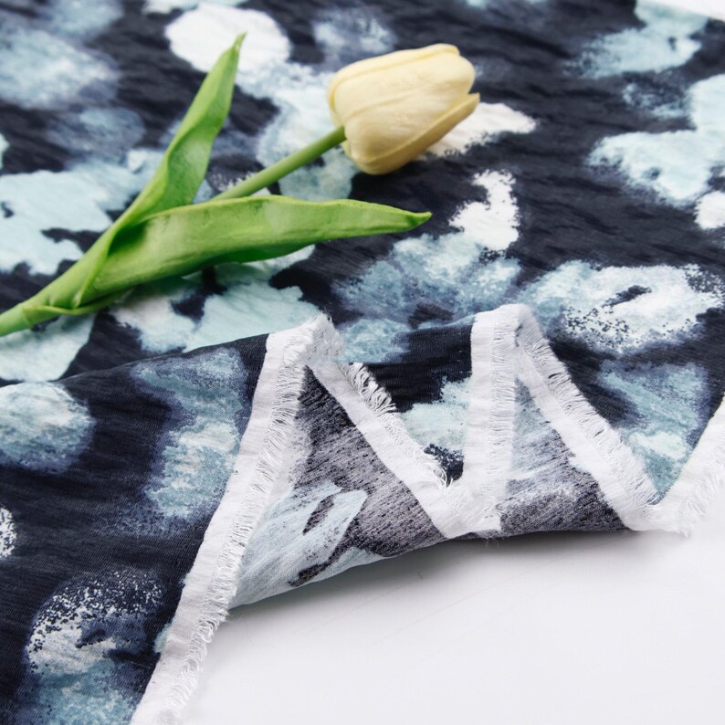 May include: Close-up of a fabric with a dark blue background and a pattern of light blue and white shapes. The fabric has a textured appearance and a frayed edge. A yellow tulip with green leaves rests on the fabric.
