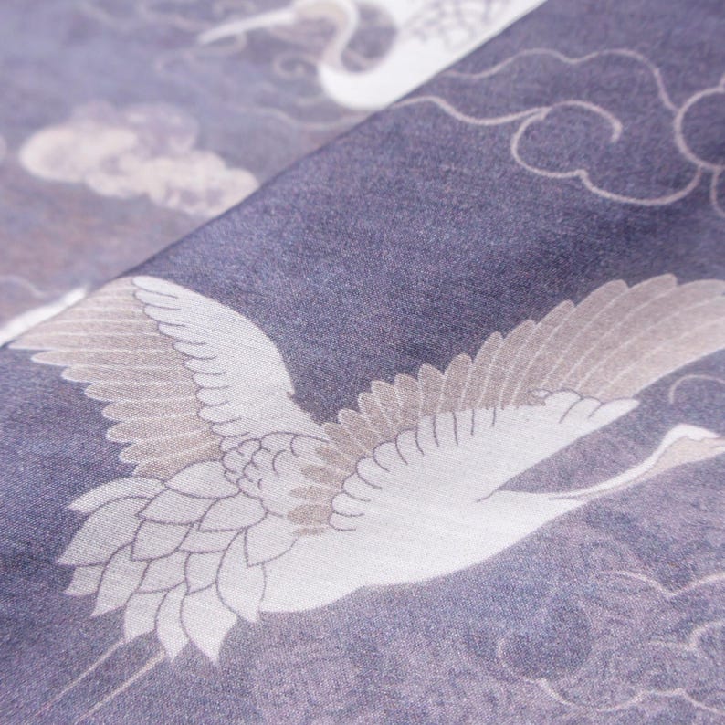 May include: Close-up of a fabric with a crane design. The fabric is a deep blue with white and tan cranes in flight, set against a background of clouds. The design is repeated across the fabric.