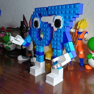 Puede incluir: Una figura de personaje pixelado azul construida con bloques de construcción entrelazados. La figura presenta una cabeza grande y cuadrada con dos ojos circulares en blanco y negro, brazos azules y manos blancas sobre dos pies blancos.