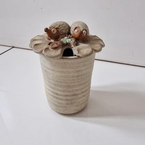 May include: A cylindrical ceramic jar with a textured, off-white finish and horizontal ridges. The lid features two sculpted brown hedgehog figures and small green accents, with a small opening for a spoon or utensil on the side of the lid.