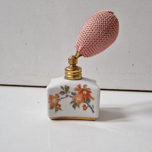 May include: A vintage perfume bottle. The white porcelain base is decorated with orange floral designs and gold accents. It has a pink, knitted atomizer. The perfume bottle is a decorative item.