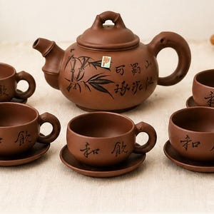 May include: A brown ceramic tea set with a teapot featuring a bamboo design and six matching cups and saucers. The teapot has a curved handle and spout. The cups and teapot have Chinese characters. The set is designed for tea ceremonies.