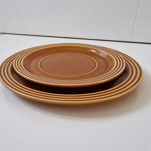 May include: Two round ceramic plates in a warm, earthy brown tone. The plates feature a series of concentric circular ridges along the outer rims, creating a textured, stepped appearance. The finish is smooth with a consistent, solid colour.