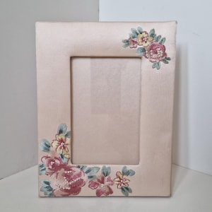 May include: A rectangular photo frame covered in pale pink satin fabric. The frame features embroidered floral motifs in shades of pink, yellow, and sage green, accented with small white beads, positioned at the top right and bottom left corners.