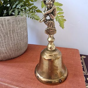 May include: A brass hand bell featuring a handle shaped like a seated creature with pointed ears. The base of the handle is inscribed with the text "CORNISH PISKY" in raised letters. The bell has a polished metallic finish with a flared bottom rim.