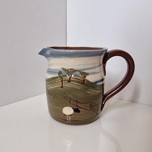 May include: A ceramic jug featuring a hand-painted landscape of green rolling hills, brown trees, and a fence. A small white sheep with a black head stands in the foreground. The jug has a dark brown handle and a blue and white sky design.