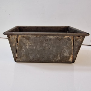 May include: A rectangular metal loaf tin with a weathered, dark grey finish. The container features tapered sides, a flat base, and a rolled rim along the top edge. The surface shows signs of oxidation and discolouration consistent with aged metal.