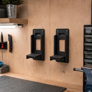 Peut inclure: Deux supports muraux noirs pour outils, avec une étagère et une ouverture rectangulaire. Ils sont fixés à un mur en bois clair. L'atelier comprend des outils, une étagère et un établi.