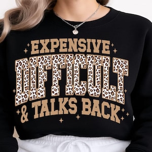 Puede incluir: Una sudadera negra de cuello redondo con el texto EXPENSIVE en letras de bloque color canela, DIFFICULT en letras de bloque con estampado de leopardo y & TALKS BACK en letras de bloque color canela. Pequeñas estrellas rodean el texto.