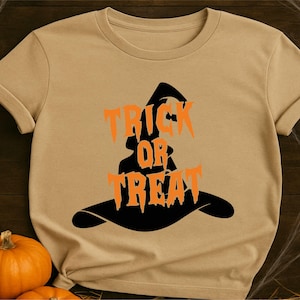 May include: A tan short-sleeved t-shirt featuring a black graphic of a witch hat. The orange text "TRICK OR TREAT" is printed in a jagged, stylized font across the center of the hat graphic, creating a bold contrast against the tan fabric.
